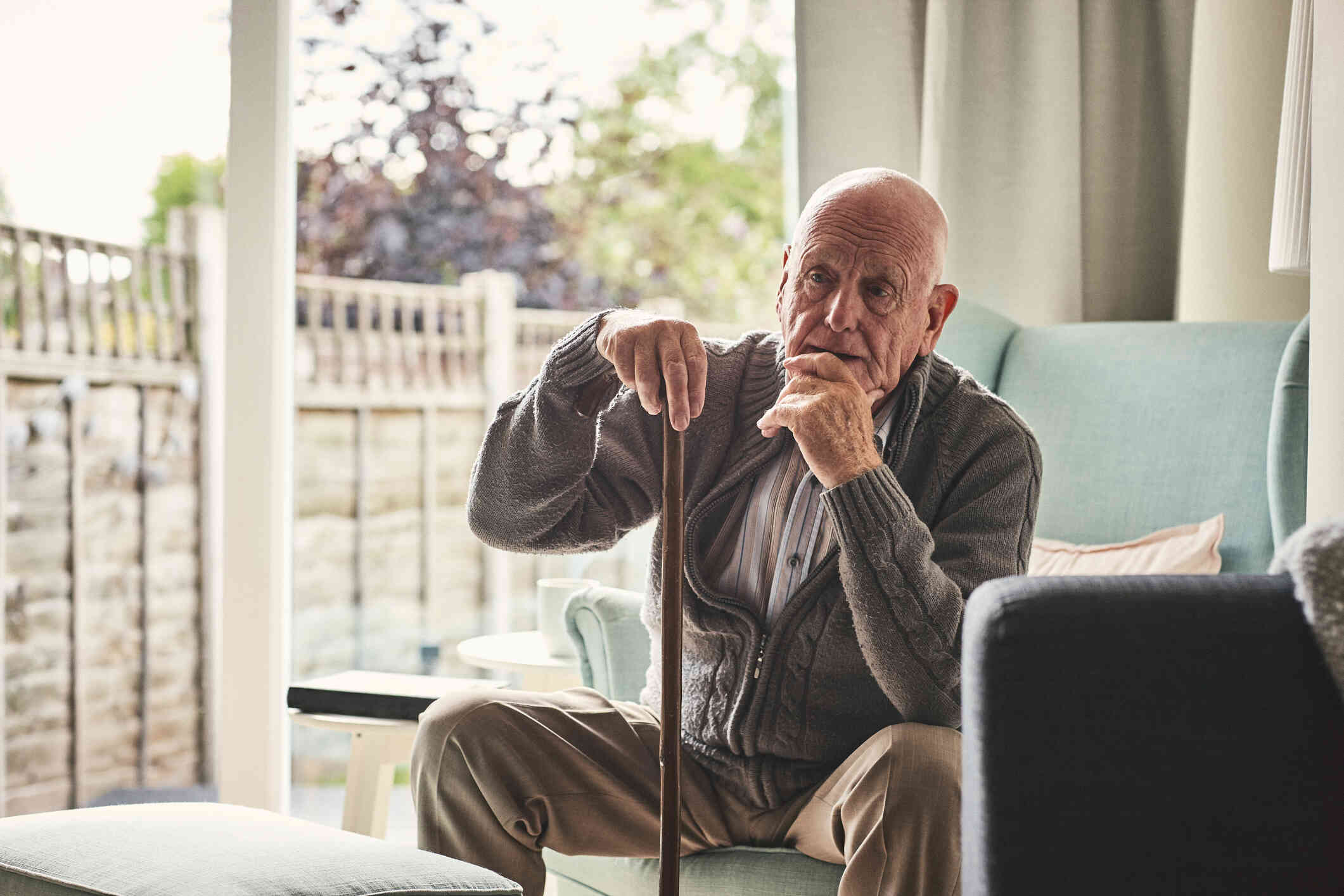 An elderly man holds a cane while sitting in an armchair in his home and gazing off with a worried expression.